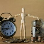 Wooden mannequin holding an alarm clock and a jar full of coins, symbolizing time and money.